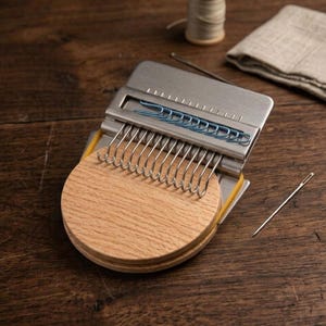 May include: A darning loom tool featuring a circular light-colored wooden base, a silver metal plate with a row of vertical hooks, and a set of blue paper clips. The device is held together by yellow rubber bands and includes a metal sewing needle.