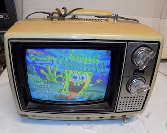 May include: A vintage Toshiba television set with a cream-colored casing. The screen displays a colorful image from the animated series "SpongeBob SquarePants." The TV has a handle, two silver knobs, and a speaker grill. The screen is approximately 10 inches.