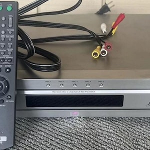 May include: A silver Sony DVD player with a 5-disc changer. The player has a black display screen and a silver remote control with the Sony logo and the word "DVD".