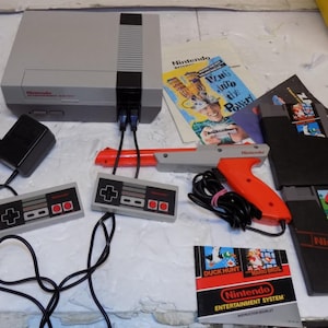 May include: A gray and black Nintendo Entertainment System console with two rectangular controllers, a red and gray light gun, a power adapter, and two game cartridges labeled Golf and Duck Hunt/Mario Bros, accompanied by two instruction booklets.