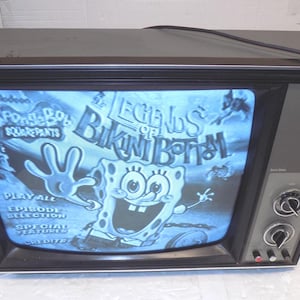May include: A vintage television displays the "Legends of Bikini Bottom" menu from Spongebob Squarepants. The TV has a black frame, gray sides, and control knobs. The screen shows a blue-toned image with cartoon characters and text.