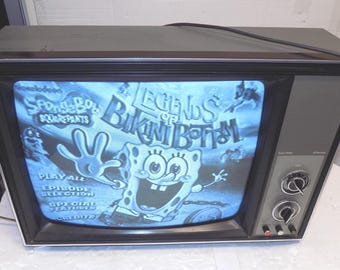 Op de afbeelding: Een vintage televisie toont het "Legends of Bikini Bottom" menu van Spongebob Squarepants. De tv heeft een zwart frame, grijze zijkanten en bedieningsknoppen. Het scherm toont een blauw getinte afbeelding met cartoonfiguren en tekst.
