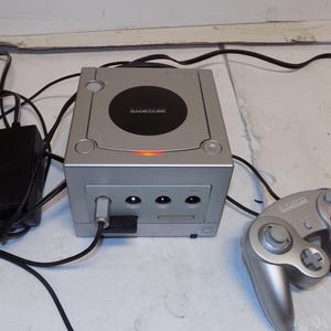 May include: A silver Nintendo GameCube video game console with a black circular disc cover, a glowing orange power light, and a matching silver controller. The console is connected to a black power adapter and includes a controller with colored buttons.