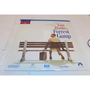 May include: A Deluxe Edition LaserDisc of the movie "Forrest Gump" starring Tom Hanks. The cover features a man sitting on a park bench with a suitcase. The title is in blue and red text.
