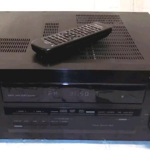 May include: Black Pioneer audio receiver with a remote control on top. The receiver has a display showing "FM 92.50". The front panel has knobs and buttons. The receiver is a home theater component.