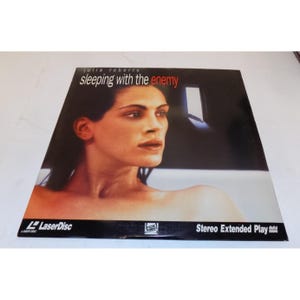 May include: LaserDisc of the movie "Sleeping with the Enemy" featuring Julia Roberts. The cover has a black background with a photo of Roberts. The title is in white and red text. The LaserDisc logo and "Stereo Extended Play" are also visible.