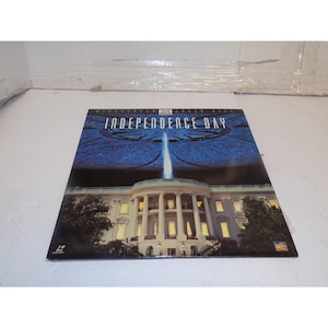 May include: A movie poster for "Independence Day" featuring the White House with a beam of light shining down from a spaceship. The top of the poster has the movie title in white text against a blue background.