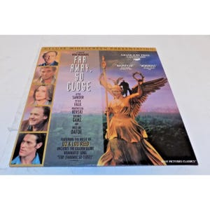 May include: A DVD case for the film "Far Away, So Close" by Wim Wenders. The cover features images of the cast, the title in large white letters, and a golden statue. The case includes text about the music by U2 & Lou Reed.