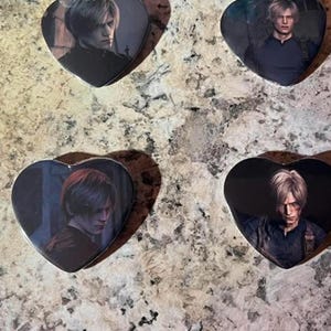 Op de afbeelding: Vier hartvormige pins met karakterportretten uit de Resident Evil videogame-serie. Elke pin toont een andere close-up afbeelding van een mannelijk personage met licht haar, geplaatst tegen een donker, gedempt achtergrondpalet.