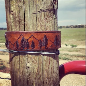 May include: A brown leather bracelet with a design of mountains and trees. The bracelet is wrapped around a wooden post.
