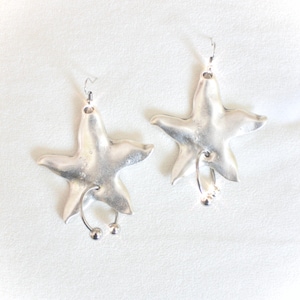 May include: A pair of silver-toned starfish earrings. Each earring features a five-pointed star shape with a curved wire and two small dangling spheres. The earrings have a polished, reflective surface and are designed to be worn with pierced ears.