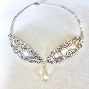 May include: A silver necklace with a large, ornate pendant and a crystal drop. The pendant is made of two pieces of metal that are joined together in the center. The metal is intricately detailed with a floral design.