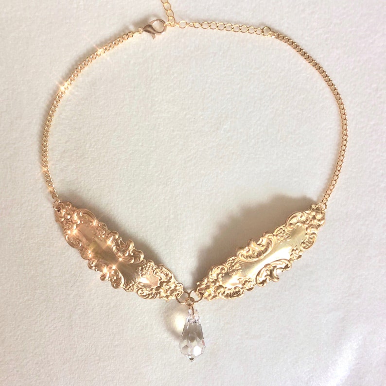 May include: A gold necklace with a delicate chain and a pendant featuring two ornate, gold, wing-shaped pieces connected by a clear crystal teardrop.