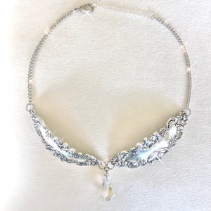 May include: A silver necklace with a pendant made from two ornate spoons and a crystal teardrop. The necklace is on a white background.