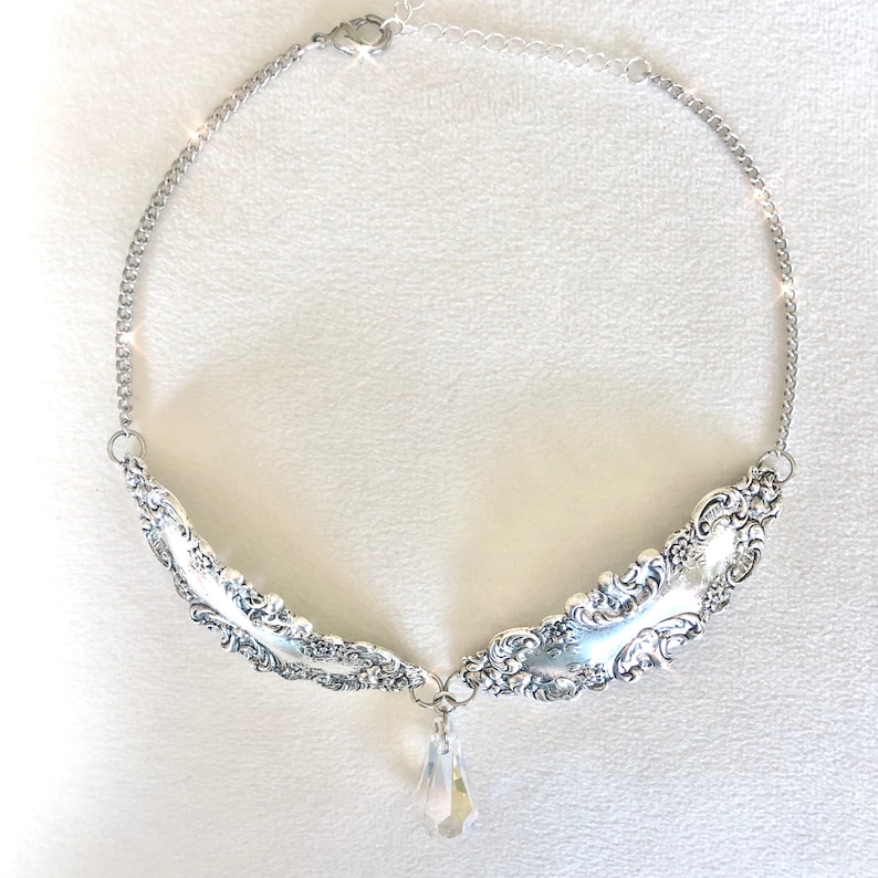 May include: A silver necklace with a pendant made from two ornate spoons and a crystal teardrop. The necklace is on a white background.