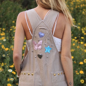 Op de afbeelding: Een beige overall met verschillende kleurrijke patches, waaronder harten, vlinders en sterren. De overall heeft gouden studs langs de onderkant.