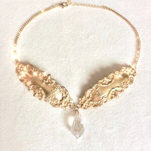 May include: A gold necklace with a large, ornate pendant featuring a crystal drop. The pendant is made of two pieces that are joined together in the center. The necklace is made of a delicate chain.