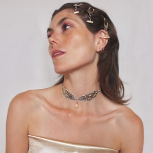 May include: A woman with long brown hair is wearing a silver choker necklace with a teardrop-shaped crystal pendant. She is also wearing several silver hair clips.