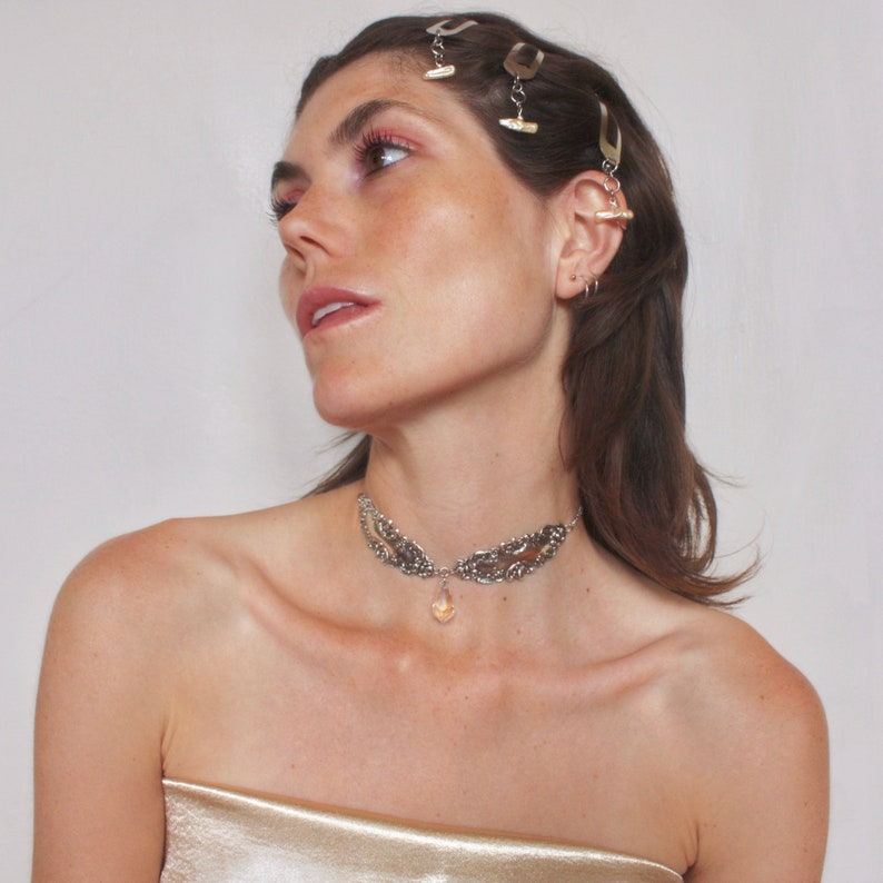 May include: A woman with long brown hair is wearing a silver choker necklace with a teardrop-shaped crystal pendant. She is also wearing several silver hair clips.