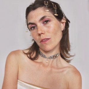 May include: A woman with long brown hair wearing a silver choker necklace with a teardrop crystal pendant. She is wearing a white strapless top and has several silver hair clips with small white stones in her hair.