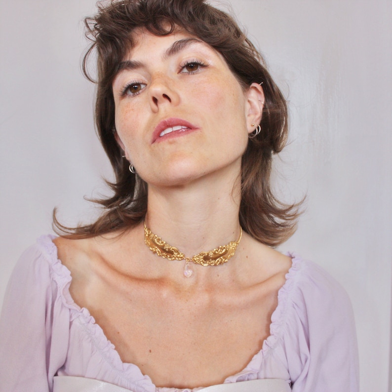 May include: A woman wearing a gold choker necklace with a pink crystal pendant. The necklace features a delicate, ornate design.