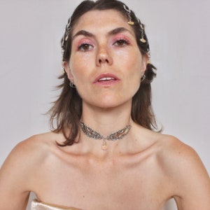 May include: A woman with brown hair wearing a silver choker necklace with a crystal pendant. She is wearing a gold top and has a silver hairpiece with small charms.
