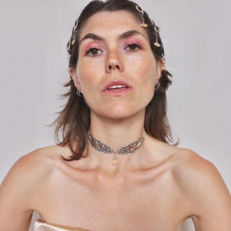 May include: A woman with brown hair wearing a silver choker necklace with a crystal pendant. She is wearing a gold top and has a silver hairpiece with small charms.