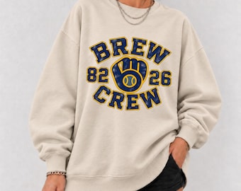 May include: A beige crewneck sweatshirt featuring a distressed navy blue and yellow graphic print that reads BREW CREW with the numbers 82 and 26 flanking a baseball glove icon in the center. The garment has long sleeves and ribbed cuffs.