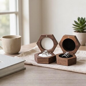 May include: Two hexagonal wooden ring boxes made of dark brown wood. The left box features a white interior lining holding a silver ring, while the right box has a black interior lining holding a silver ring. Both boxes are open and display the rings.