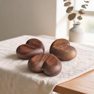 Può includere: Tre oggetti lisci e levigati a forma di cuore, intagliati in legno marrone scuro con venature visibili. Ogni cuore presenta una forma arrotondata e organica con una distinta fessura sulla parte superiore, che mette in risalto la texture naturale.