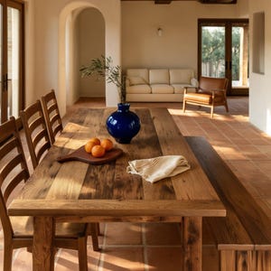 May include: A rectangular wooden dining table with a dark blue ceramic vase containing a leafy branch, a wooden cutting board with three oranges, and a folded beige cloth napkin placed on the surface, surrounded by wooden chairs and a bench.
