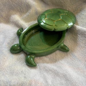 Pode incluir: Uma caixa de bugigangas em forma de tartaruga verde com tampa removível. O corpo da tartaruga é de um verde profundo e cintilante com um acabamento liso. A tampa em forma de carapaça apresenta um padrão segmentado delineado em dourado.