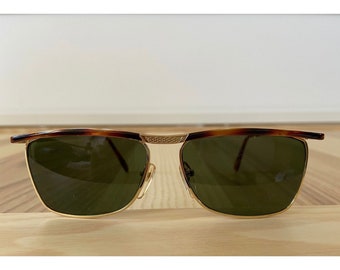 May include: Gold-framed sunglasses with green lenses. The top of the frame features a tortoiseshell pattern. The sunglasses have a classic, rectangular shape and are set on a wooden surface.