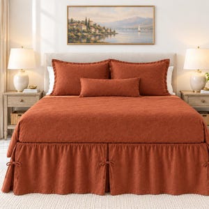 May include: A rust-colored bedding set featuring a textured quilt, two matching standard pillow shams, one rectangular bolster pillow, and a bed skirt with decorative side ties, all displayed on a bed with a neutral headboard.