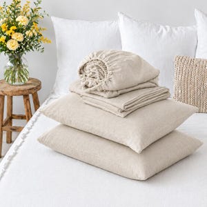 May include: A set of beige linen bedding including two rectangular pillows stacked on top of each other, a folded flat sheet, and a fitted sheet with elastic edges. The fabric has a visible woven texture in a neutral light tan color.