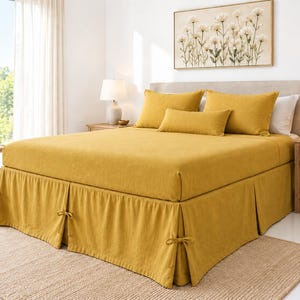 May include: A mustard yellow bed skirt featuring a pleated design with decorative fabric ties at each corner and along the sides. The bed skirt is made of a solid-colored textile and is positioned beneath a matching mustard yellow bed sheet.