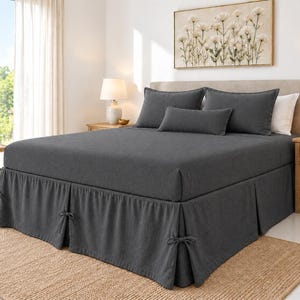 May include: A dark grey bed skirt featuring a pleated design with decorative fabric ties at each corner and along the sides. The bed skirt is made of a solid-coloured fabric and is positioned beneath a mattress with matching dark grey bedding.