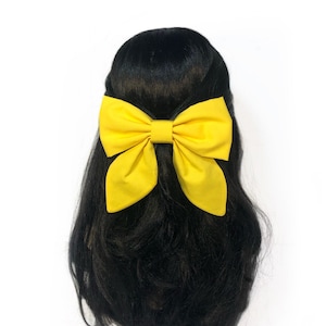 May include: A large yellow bow hair clip. The bow is made of fabric and is tied in a classic bow shape.