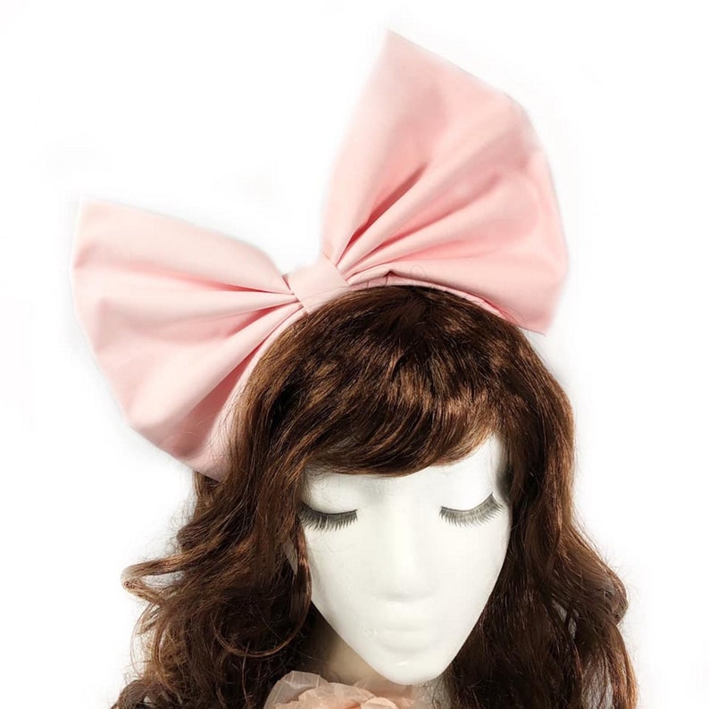 May include: A large pink fabric bow headband. The headband is worn on a mannequin with brown hair.
