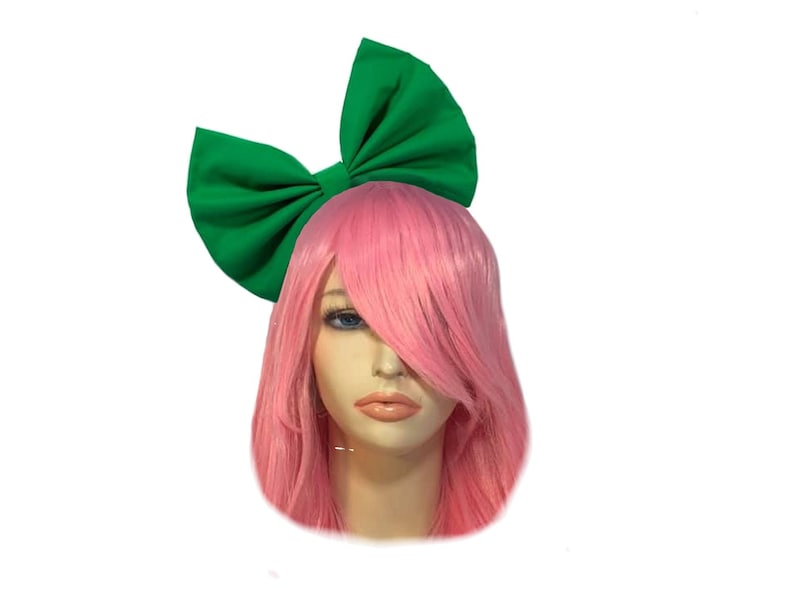 May include: A large green bow headband with a pink wig. The bow is made of fabric and is tied in a large knot. The wig is long and straight.