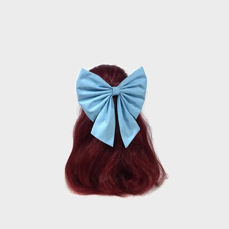 May include: A large, sky blue fabric bow tied in a simple knot. The bow is worn in a ponytail on a model with long, dark red hair.