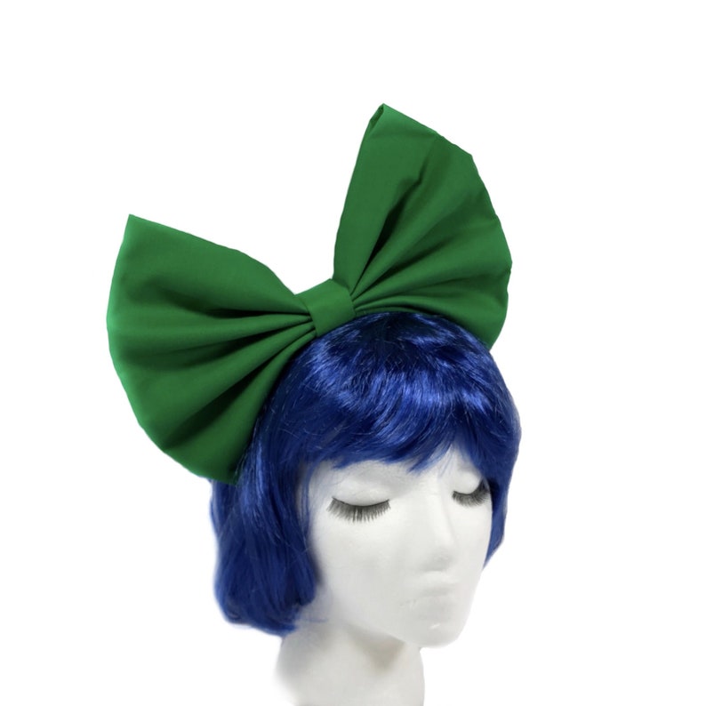 May include: A large green fabric bow headband. The bow is tied in a large, oversized style. The headband is worn on a mannequin with blue hair.