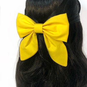 May include: A large yellow bow made of fabric. The bow is tied in a classic style with a center loop and two long tails. The bow is worn in the hair of a person with long black hair.