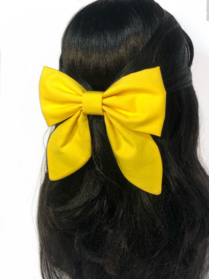 May include: A large yellow bow made of fabric. The bow is tied in a classic style with a center loop and two long tails. The bow is worn in the hair of a person with long black hair.