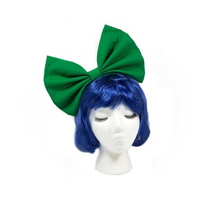 May include: A large green fabric bow headband. The bow is tied in a large, dramatic loop. The headband is worn on a mannequin with blue hair.