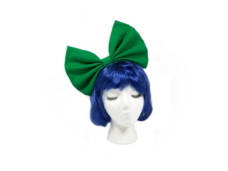 May include: A large green fabric bow headband. The bow is tied in a large, dramatic loop. The headband is worn on a mannequin with blue hair.