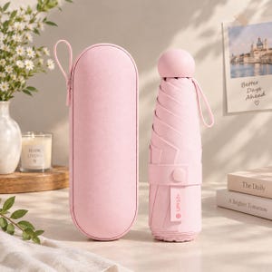 May include: A light pink compact umbrella with a matching zippered carrying case. The umbrella features a rounded handle, a wrist strap, and a fabric strap with a snap button closure labeled UPF50+. The exterior is a solid, matte light pink.
