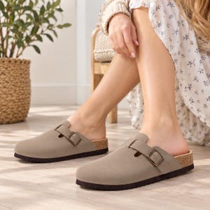 May include: A pair of taupe-colored slip-on clogs featuring a rounded toe, a single adjustable strap across the instep with a metallic buckle, a cork-textured midsole, and a dark brown textured outsole for traction and stability.