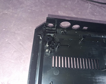 May include: A black plastic electronic device housing featuring a series of vertical ventilation slots, internal screw mounting posts, and circular port openings along the top edge of the rectangular frame.