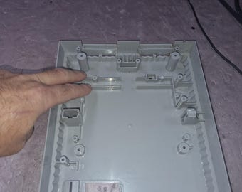 May include: A light gray plastic interior shell of a video game console, featuring molded screw posts, internal support ribs, and rectangular port openings. The structure is rectangular with a hollow center and raised mounting points.
